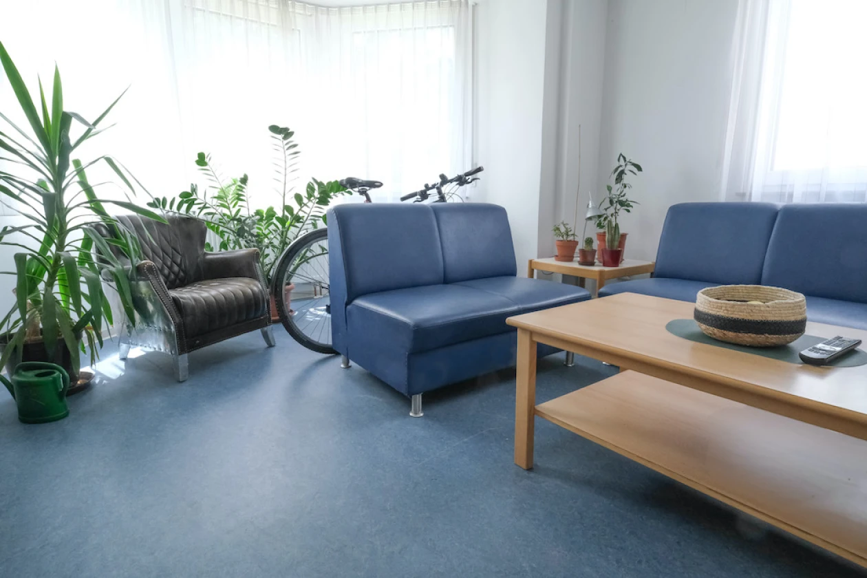 Blick auf eine Sitzecke mit zwei blauen Sofas, einem braunen Sessel und Holztisch. Gr&uuml;npflanzen und ein Fahrrad stehen im Hintergrund. Fenster mit wei&szlig;en Vorh&auml;ngen sorgen f&uuml;r eine helle und freundliche Atmosph&auml;re.