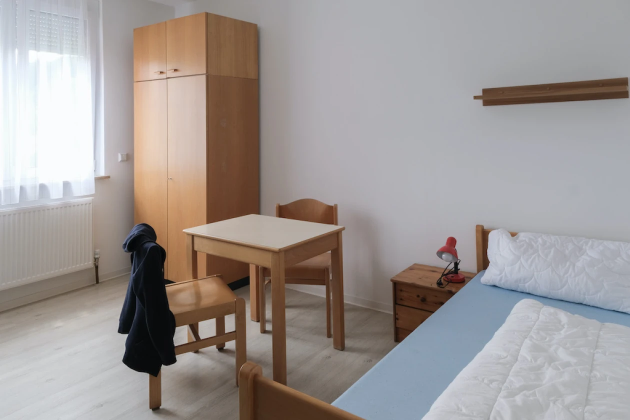 Ein Zimmer mit wei&szlig;en W&auml;nden und hellem Boden mit Holzschrank, kleinem Holztisch mit zwei Holzst&uuml;hlen sowie ein Bett mit blauem Bettbezug, wei&szlig;er Decke und wei&szlig;em Kissen. Eine Jacke h&auml;ngt an &uuml;ber der Stuhllehne. Ein Fenster mit wei&szlig;em Vorhang l&auml;sst Tageslicht in den Raum.