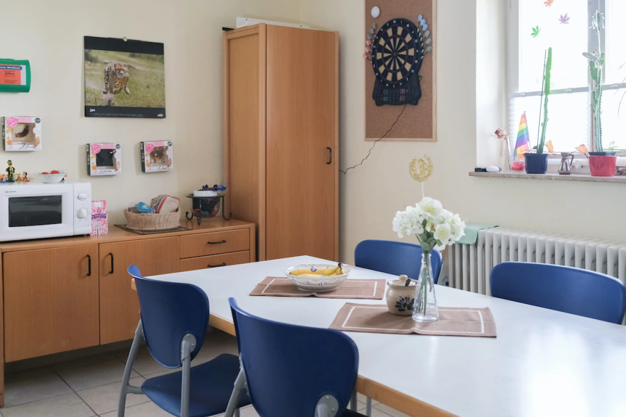Ein Raum mit wei&szlig;em Tisch, blauen St&uuml;hlen, Holzschrank, Mikrowelle und Dartscheibe an der Wand. Wei&szlig;e Blumen in einer Vase auf dem Tisch, Fenster mit Topfpflanzen im Hintergrund.