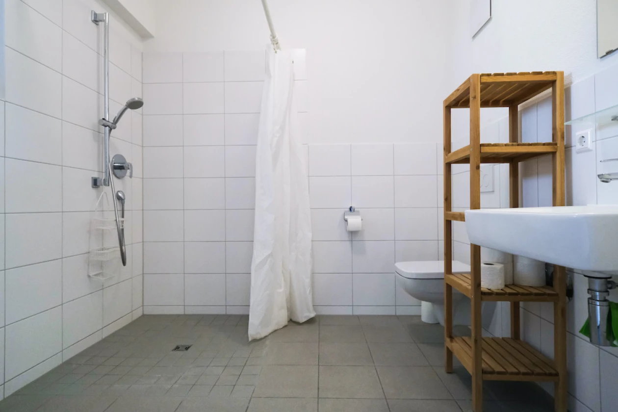 Helles Badezimmer mit bodenebener Dusche, wei&szlig;em Duschvorhang, Toilette und Holzregal. Das Bad hat graue Bodenfliesen und wei&szlig;e Wandfliesen.