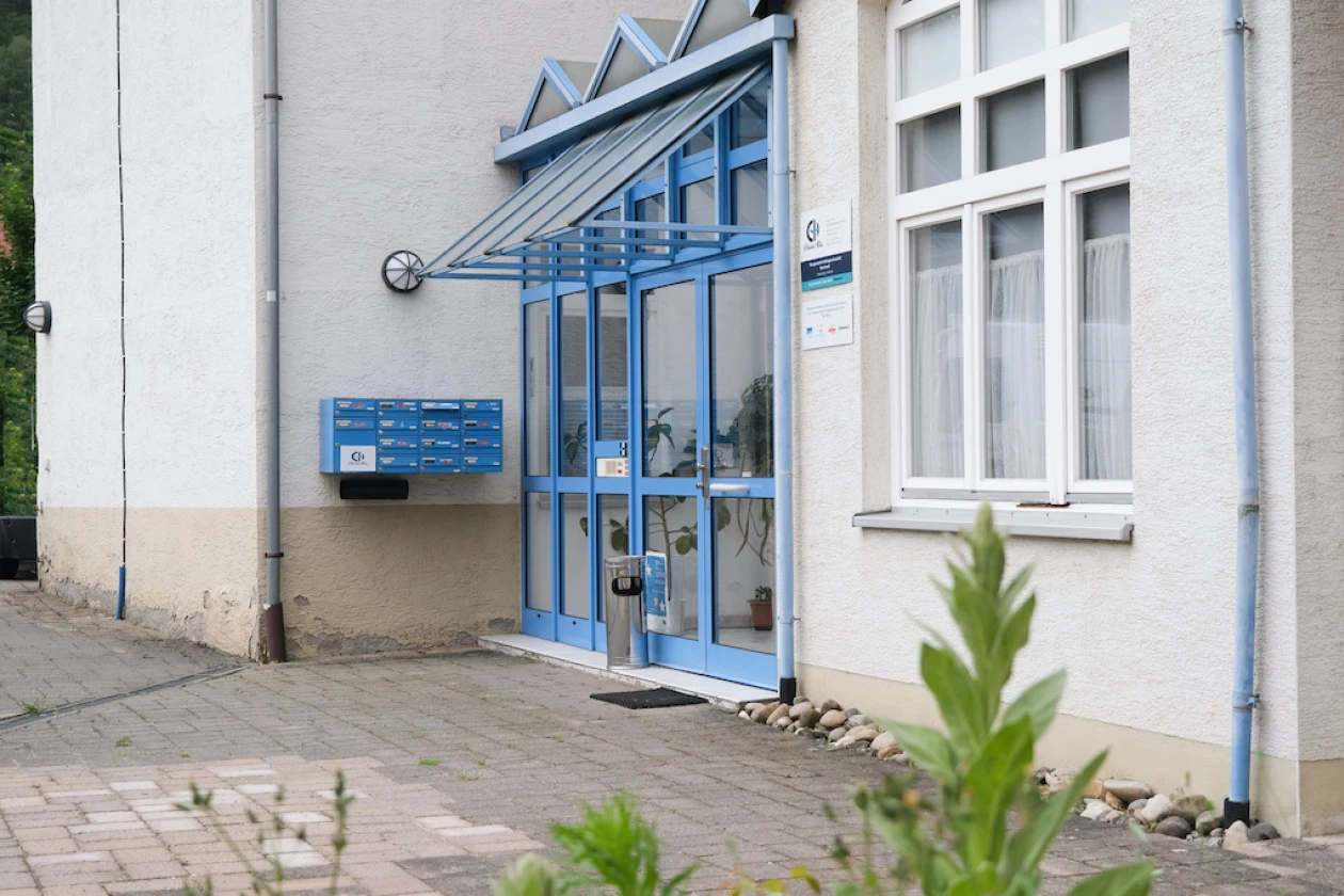 Eingangsbereich eines Geb&auml;udes mit Briefkastenanlage. Haust&uuml;re und Briefkastenanlage sind in blauer Farbe. Neben der Eingangst&uuml;re h&auml;ngt ein Schild mit dem Logo der Erlacher H&ouml;he.