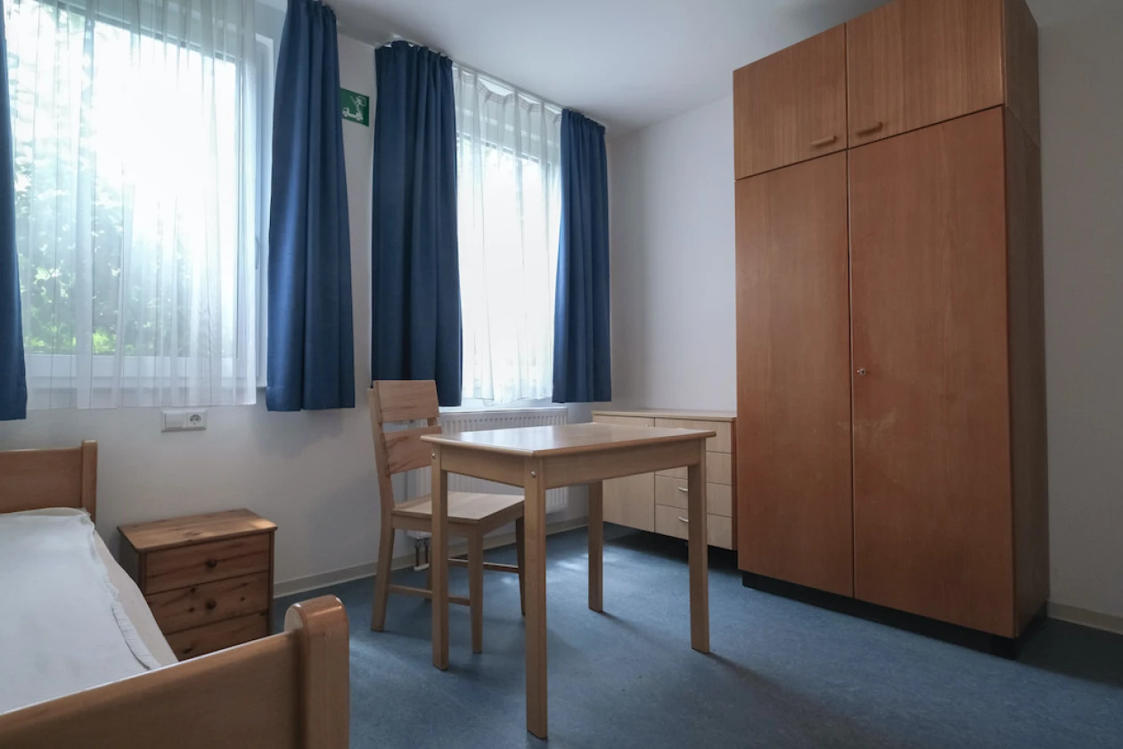 Ein Raum mit Einzelbett, ein Holztisch mit Stuhl, ein gro&szlig;er Holzschrank und blauen Vorh&auml;ngen an den Fenstern. Der Raum hat wei&szlig;e W&auml;nde und einen blauen Boden.