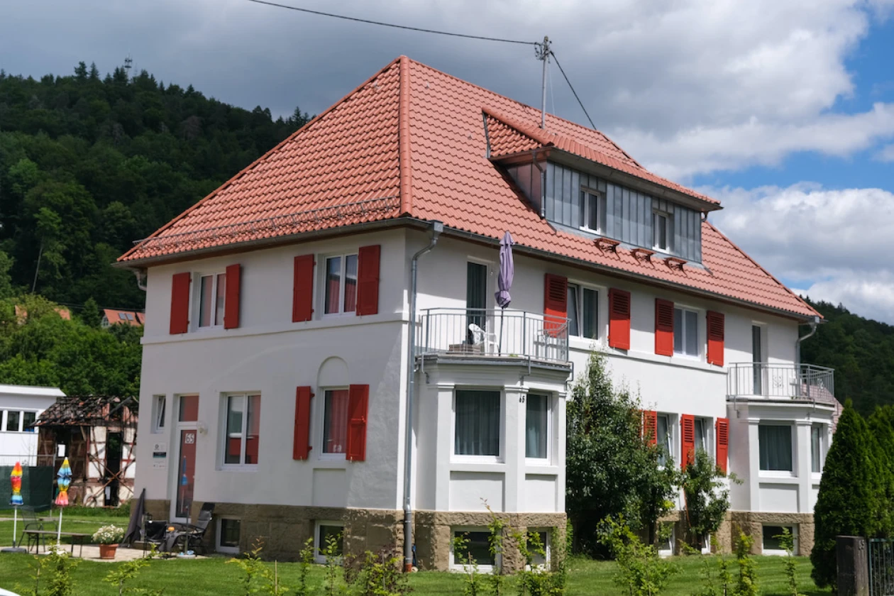 Renovierte Villa mit wei&szlig;er Fassade und roten Fensterl&auml;den in einem gepflegten Gartengrundst&uuml;ck. Links und rechts sind zwei Balkone auf kleinen turmartigen Anbauten.
