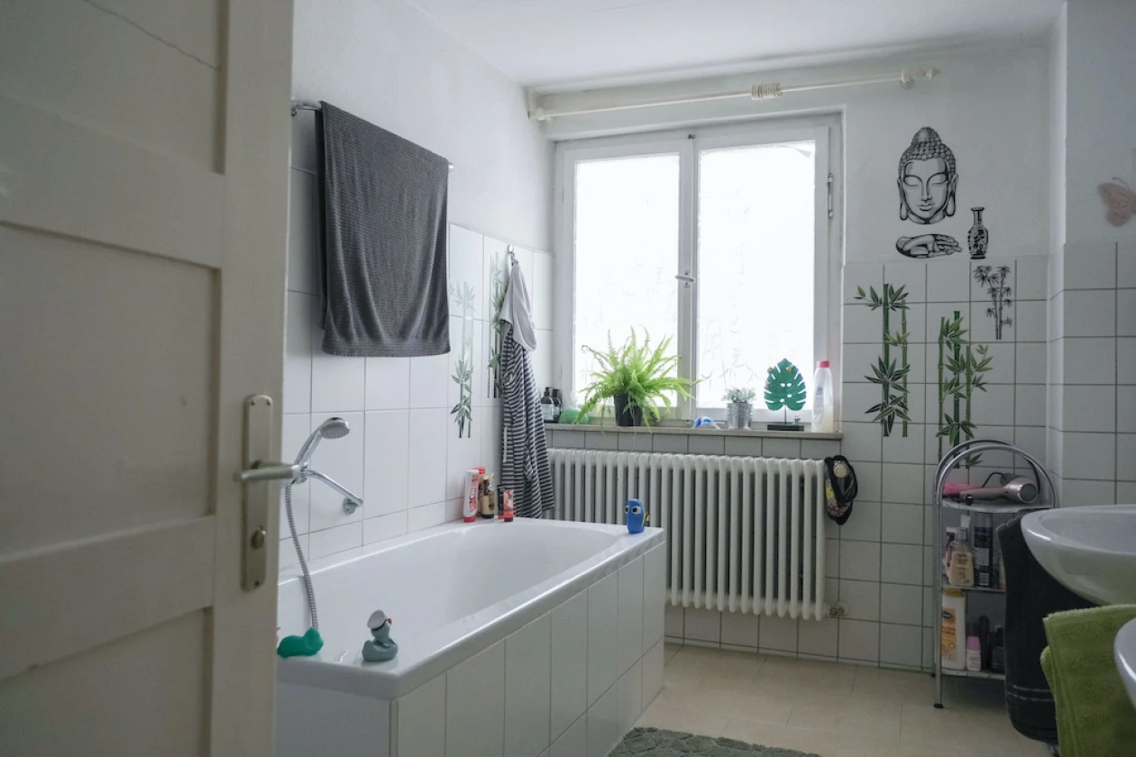 Helles, modernes Badezimmer mit wei&szlig;en Fliesen, wei&szlig;er Badewanne, Waschbecken, Fenster mit Pflanzen, grauen Handt&uuml;chern und dekorativen Wandzeichnungen im asiatischen Stil.