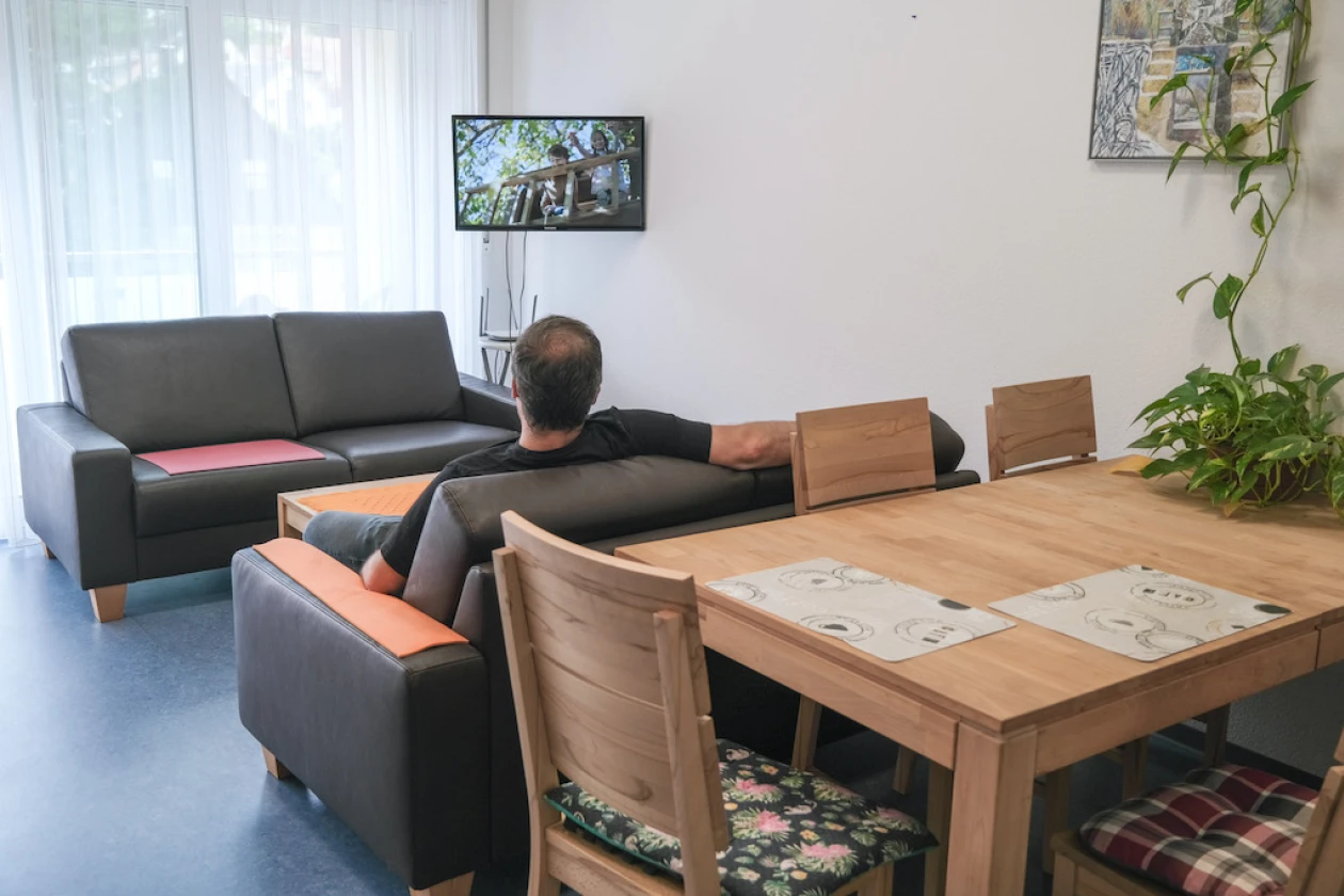 Ein Raum mit grauem Sofa, Holztisch und St&uuml;hlen. Ein Mann sitzt auf dem Sofa und schaut auf einen Fernseher an der Wand. Eine Zimmerpflanze und ein Kunstwerk vervollst&auml;ndigen die gem&uuml;tliche Einrichtung.