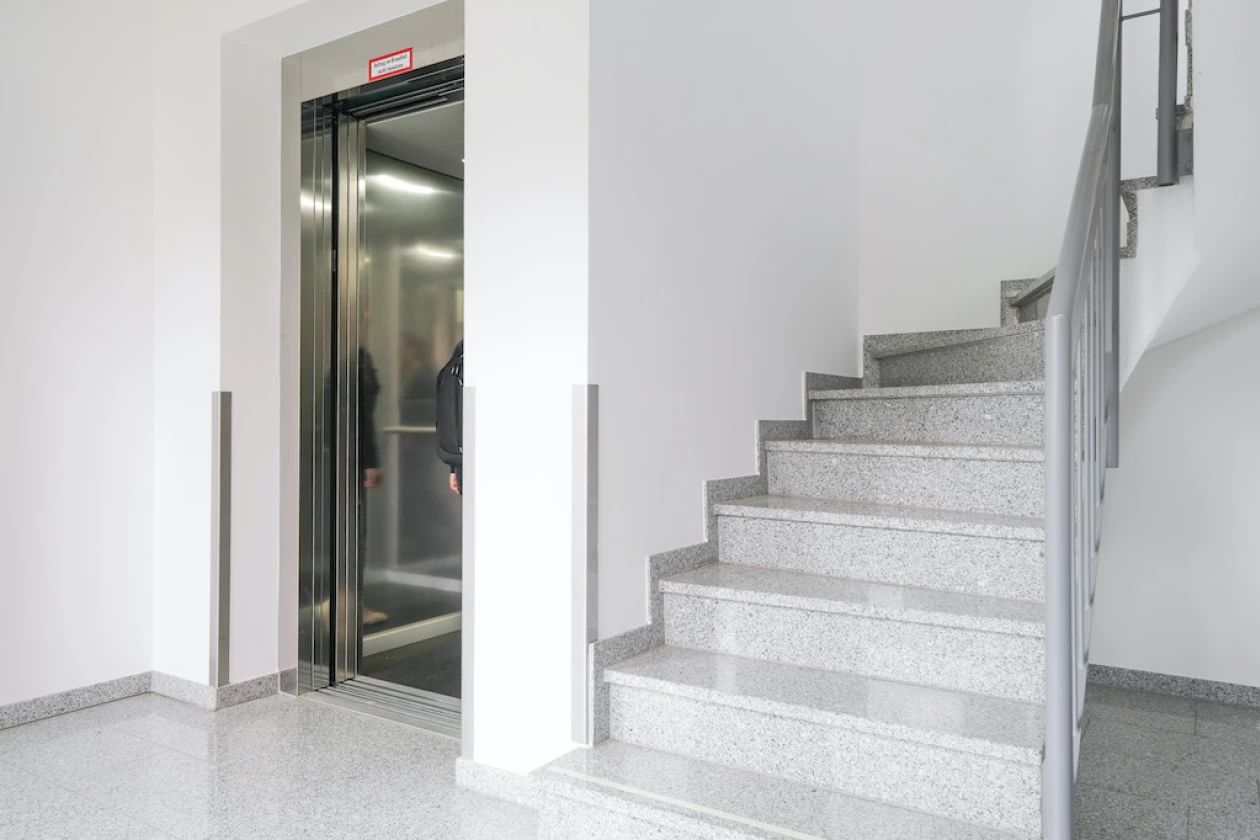 Ein modernes Treppenhaus. Der Aufzug befindet sich neben einer grauen Steintreppe in einem hellen, gepflegten Eingangsbereich mit wei&szlig;en W&auml;nden und einem gl&auml;nzenden Boden.