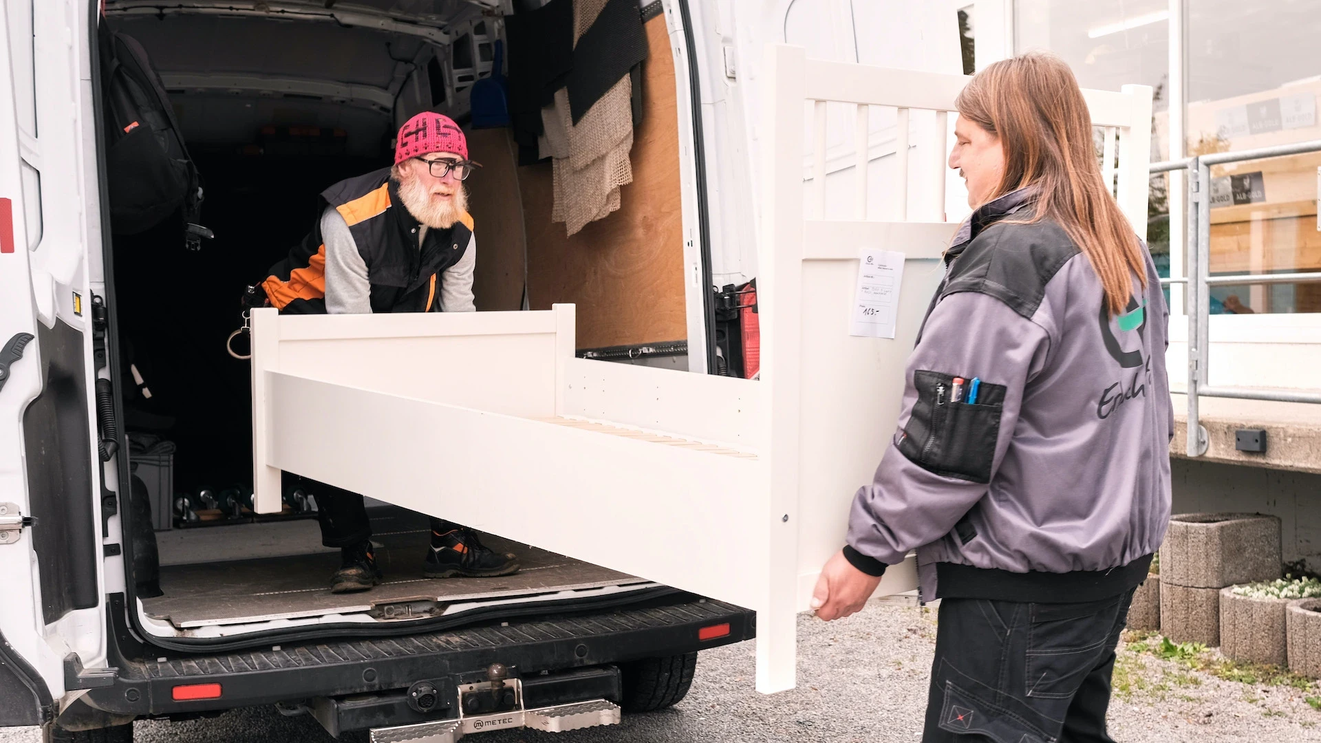 Zwei Personen laden ein wei&szlig;es M&ouml;belst&uuml;ck aus einem wei&szlig;en Transporter, eine Person steht im Fahrzeug, die andere davor auf dem Hof.