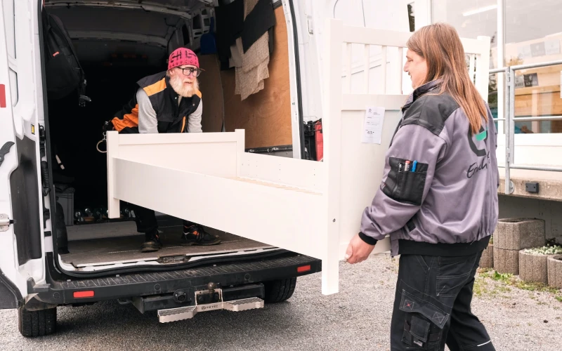 Zwei Personen laden ein wei&szlig;es M&ouml;belst&uuml;ck aus einem wei&szlig;en Transporter, eine Person steht im Fahrzeug, die andere davor auf dem Hof.