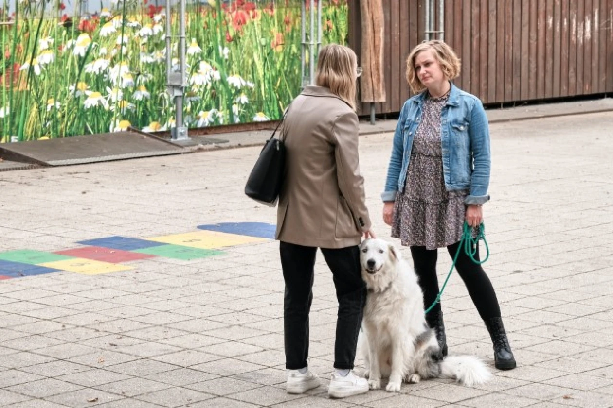 Zwei Frauen unterhalten sich auf einer gepflasterten Hoffl&auml;che. Zwischen ihnen sitzt ein wei&szlig;er Hund. Im Hintergrund ist eine Holzwand, eine Fassade mit Blumenwiesendekor und ein buntes Bodenmuster erkennbar.