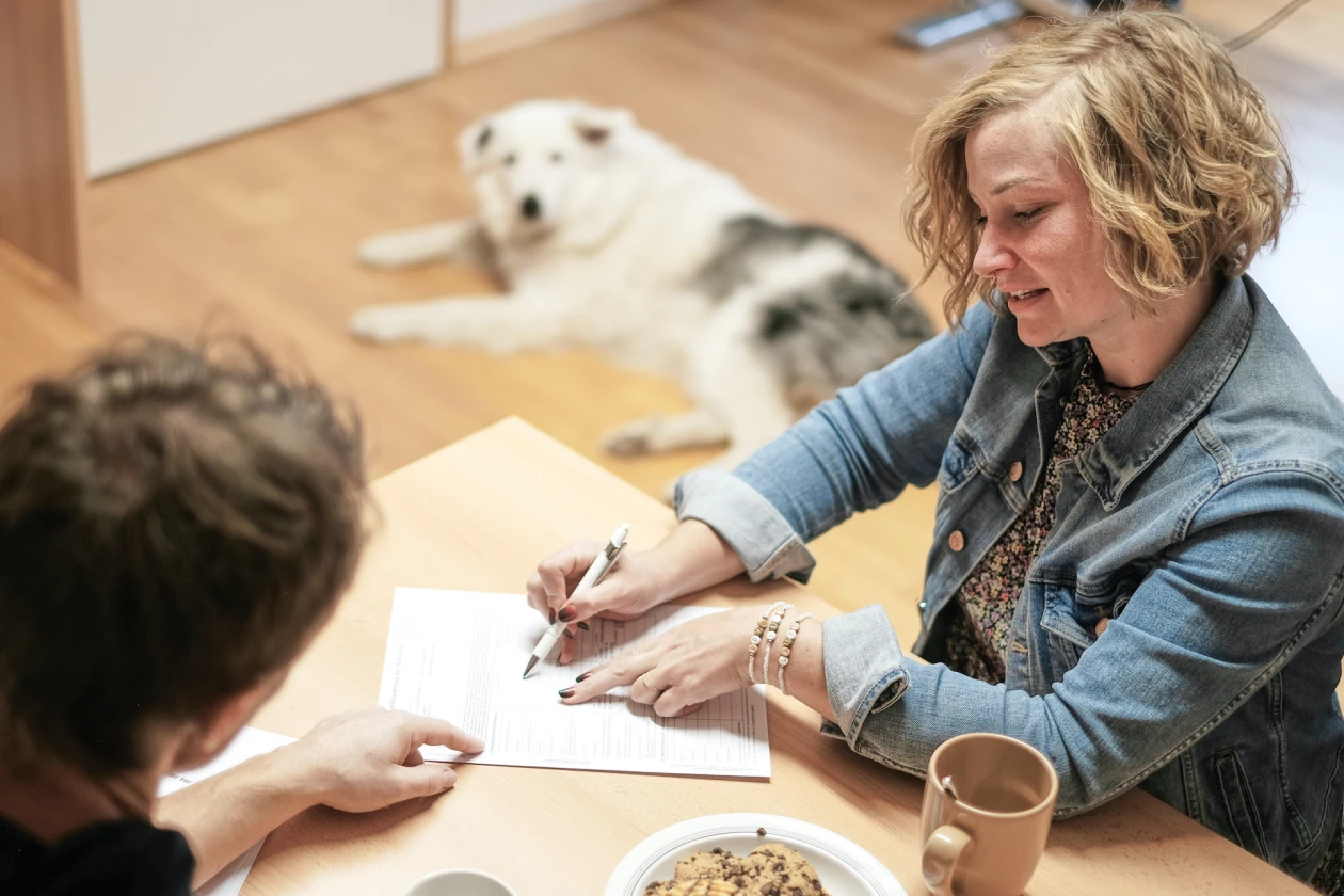 Zwei Personen sitzen an einem Tisch. Die Frau in der Jeansjacke deutet mit dem Kugelschreiber auf eine Textpassage auf einem Dokument, die andere Person sitzt mit dem R&uuml;cken zur Kamera und h&ouml;rt zu. Im Hintergrund liegt ein wei&szlig;er Hund auf dem Boden. Auf dem Tisch steht eine Tasse und eine Sch&uuml;ssel mit Keksen.