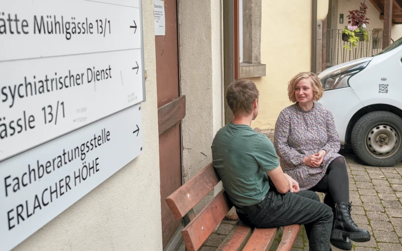Zwei Personen sitzen auf einer Bank vor einem Geb&auml;ude. Im Hintergrund ist ein Fahrzeug zu sehen. Im Vordergrund sind Hinweisschilder zum psychiatrischen Dienst und zur Fachberatungsstelle Erlacher H&ouml;he zu sehen.