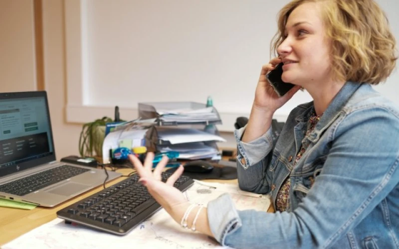 Junge Frau mit blonden gewellten Haaren und Jeansjacke telefoniert am Smartphone, w&auml;hrend sie an ihrem Arbeitsplatz mit Laptop, Tastatur und Dokumentenablage sitzt.
