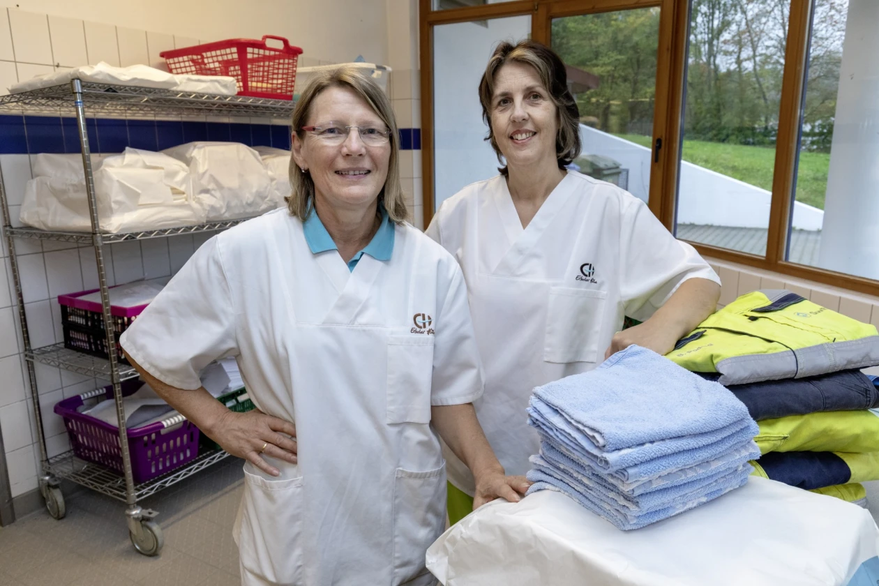 Zwei Frauen in wei&szlig;en Berufskleidung stehen in einem W&auml;scheraum mit Regalen, K&ouml;rben und Handtuchstapeln und l&auml;cheln in die Kamera. Sie stehen vor einem Fenster mit Blick ins Gr&uuml;ne.