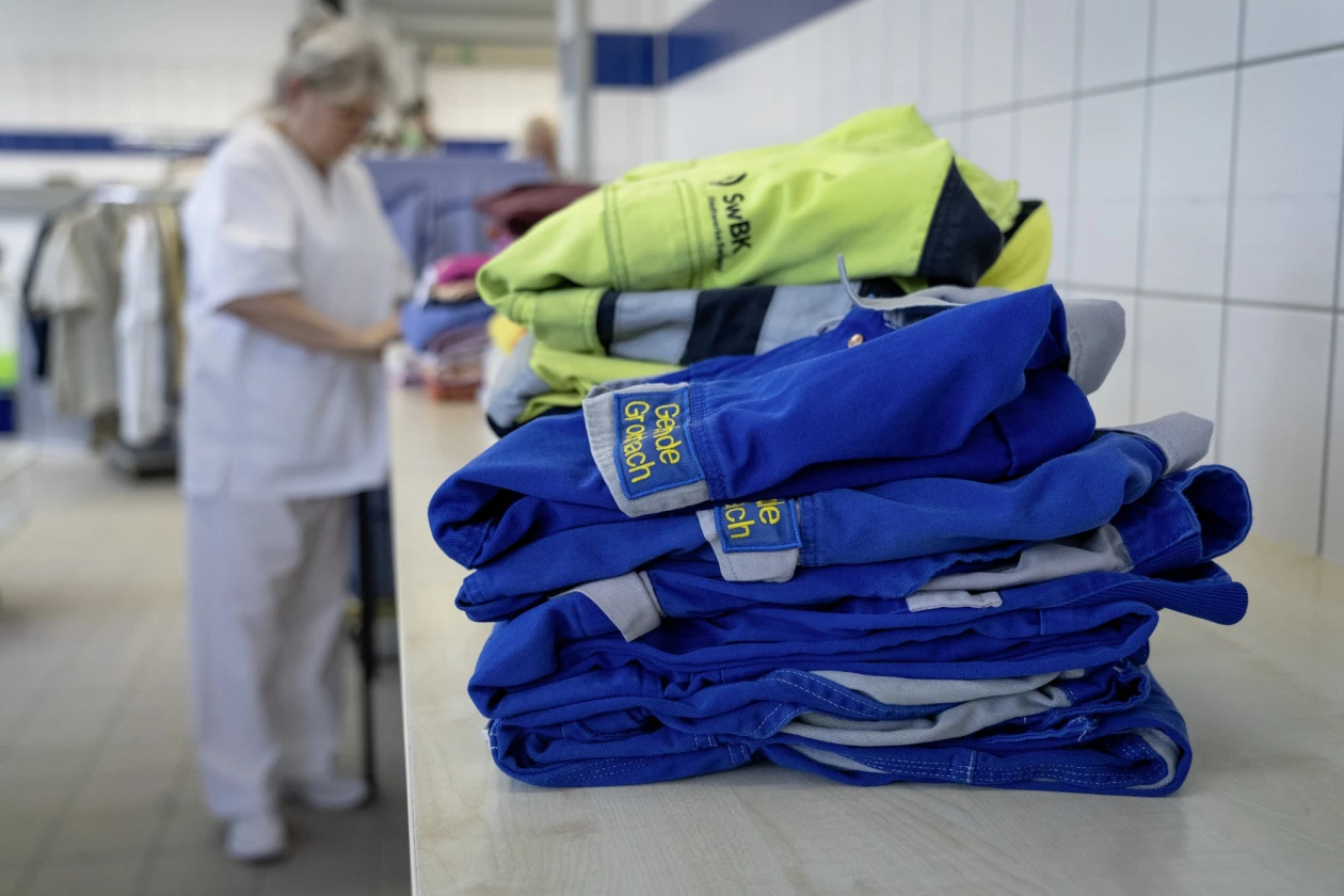 Gestapelte blaue und gelbe Arbeitskleidung im Vordergrund, im Hintergrund verschwommen eine Person in wei&szlig;er Arbeitskleidung.