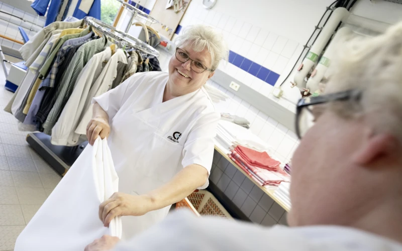 Eine Frau in wei&szlig;er Berufskleidung in einer W&auml;scherei l&auml;chelt in die Kamera. Sie legt zusammen mit einer weiteren Person im Vordergrund ein wei&szlig;es Textilst&uuml;ck zusammen. Im Hintergrund h&auml;ngen Kleidungst&uuml;cke auf einem Kleiderst&auml;nder.