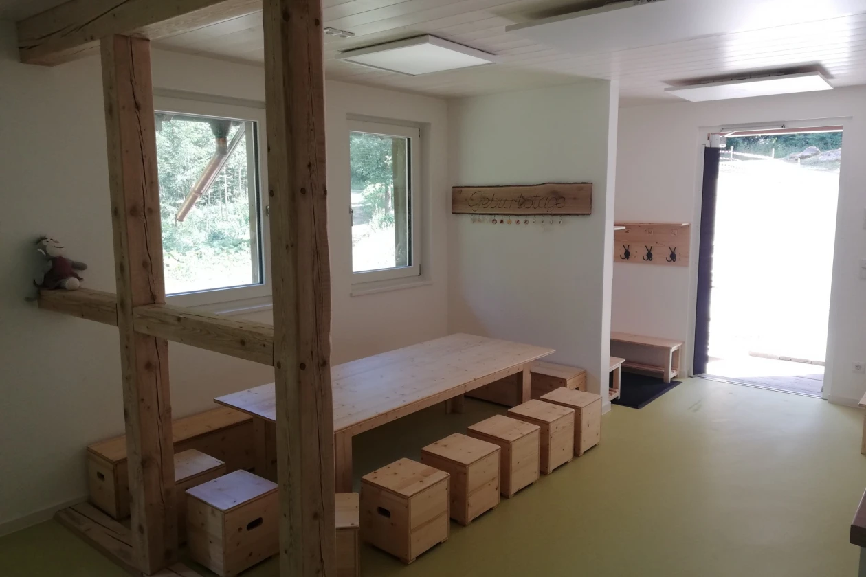 Raum ist mit einem Holztisch, sichtbaren Balken, einer Sitzbank und w&uuml;rfelf&ouml;rmigen Holzhockern ausgestattet. Zwei Fenster und eine ge&ouml;ffnete Eingangst&uuml;r sorgen f&uuml;r Helligkeit.