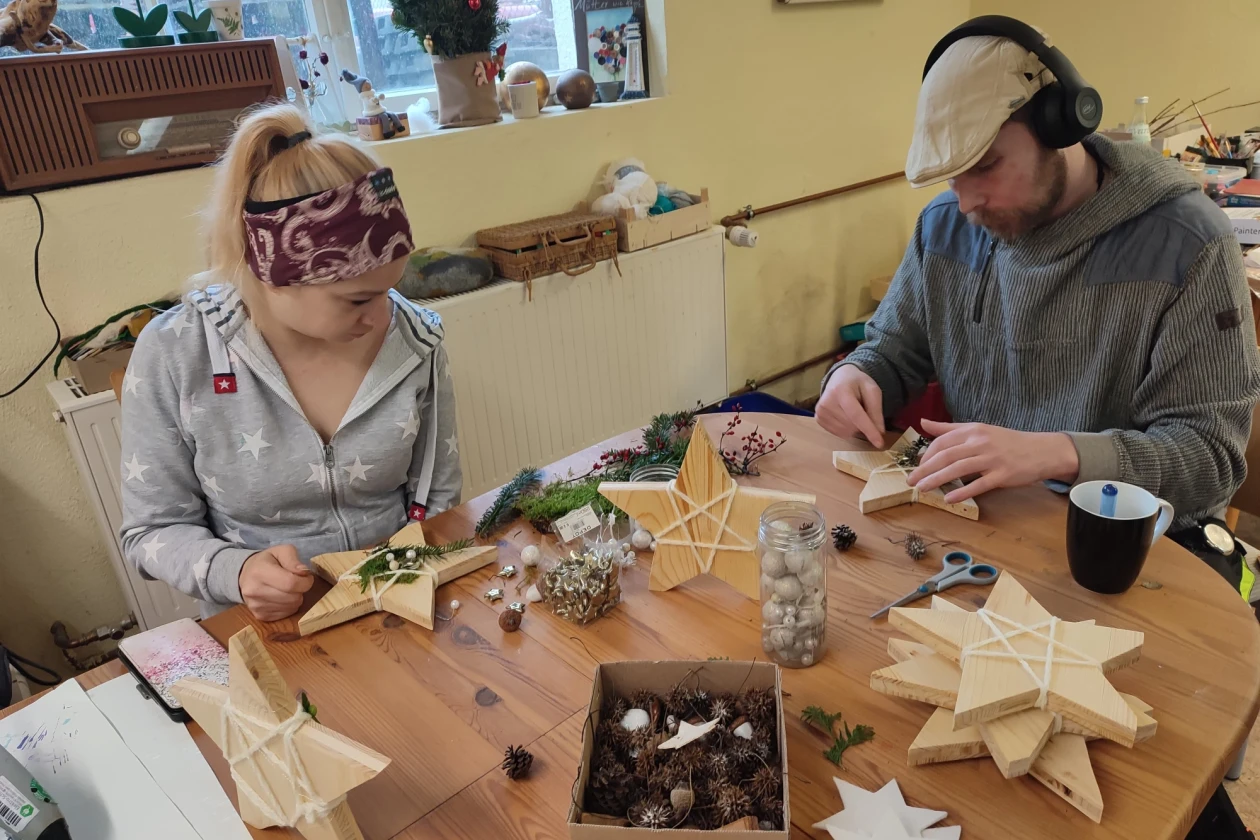 Eine Frau und ein Mann sitzen an einem Tisch und bekleben Holzsterne mit verschiedenen Bastelmaterialien.