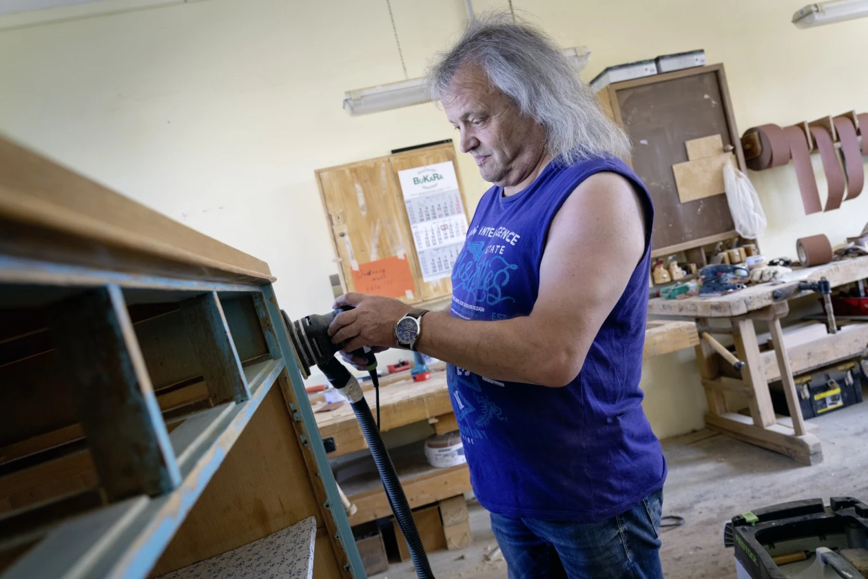 Ein Mann mit langem grauem Haar in blauem Top schleift in einer Werkstatt die Oberfl&auml;che eines Schranks ab. Im Hintergrund ist Werkzeug, Werkbank und Werkstattausstattung zu sehen.