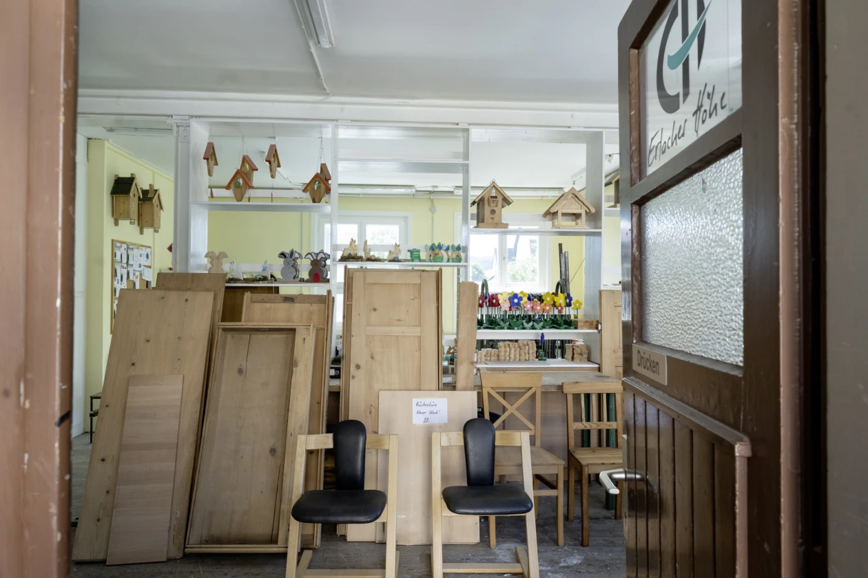 Blick in eine Holzwerkstatt durch die offene Eingangst&uuml;re. Es stehen Holzt&uuml;ren, Bretter und St&uuml;hle im Raum. Im Regal im Hintergrund stehen Vogelh&auml;uschen und Meisenkn&ouml;del-H&auml;uschen h&auml;ngen daneben.