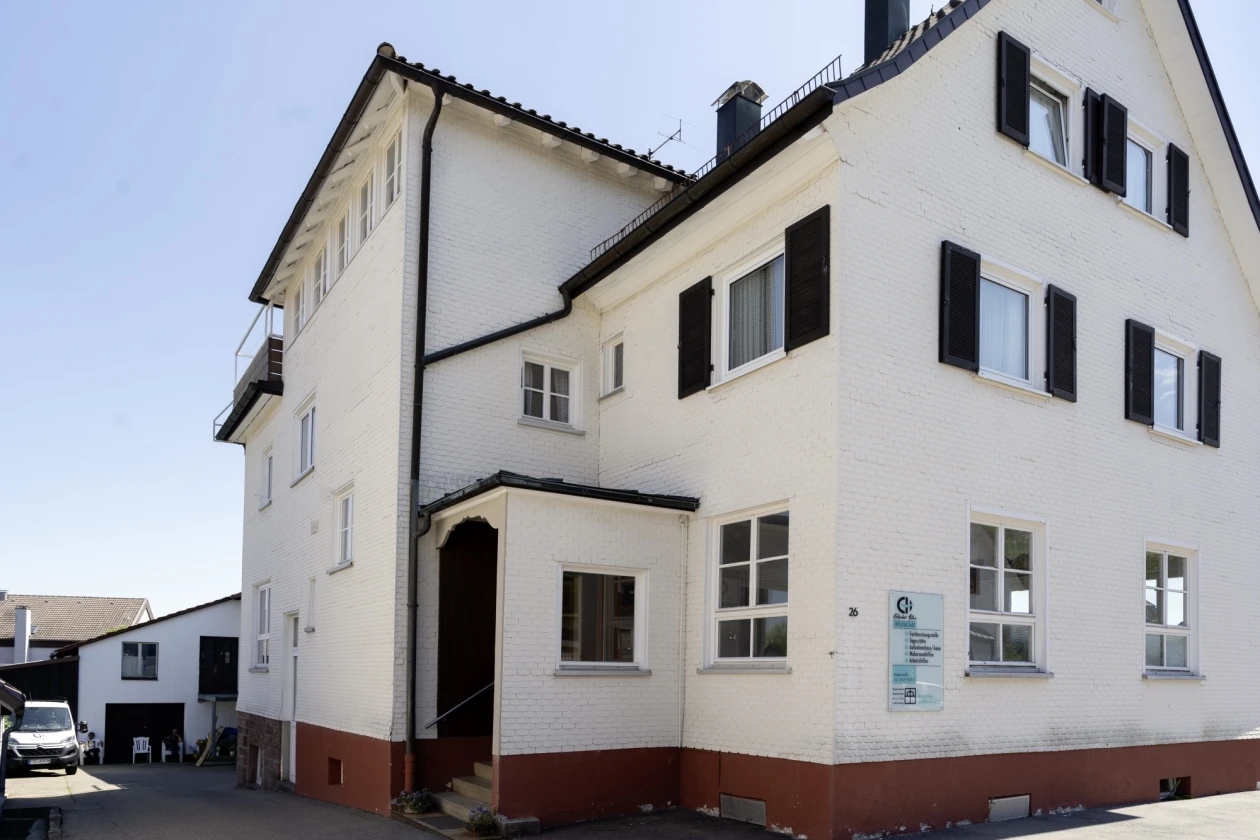 Ein wei&szlig;es, mehrst&ouml;ckiges Haus mit der Beschilderung der Erlacher H&ouml;he an der Fassade. Schwarze Fensterl&auml;den und ein rotbrauner Sockel setzen farbige Akzente. Im Hintergrund ist ein Fahrzeug und ein weiteres Geb&auml;ude zu sehen.