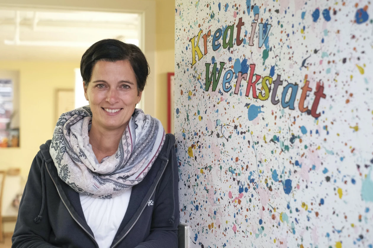 L&auml;chelnde Frau in gemustertem Schal und schwarzer Jacke steht vor einer bunten Wand mit der Aufschrift "Kreativ Werkstatt".
