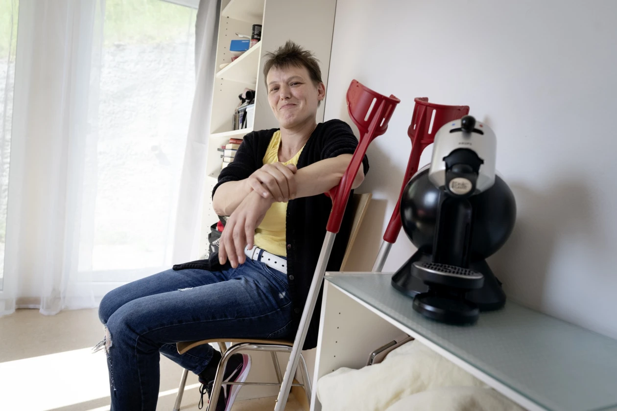 Eine Frau sitzt l&auml;chelnd auf einem Stuhl, neben sich zwei rote Kr&uuml;cken, links von ihr ein hohes wei&szlig;es Regal, rechts von ihr steht eine Kaffeemaschine auf einem kleinen Regal.