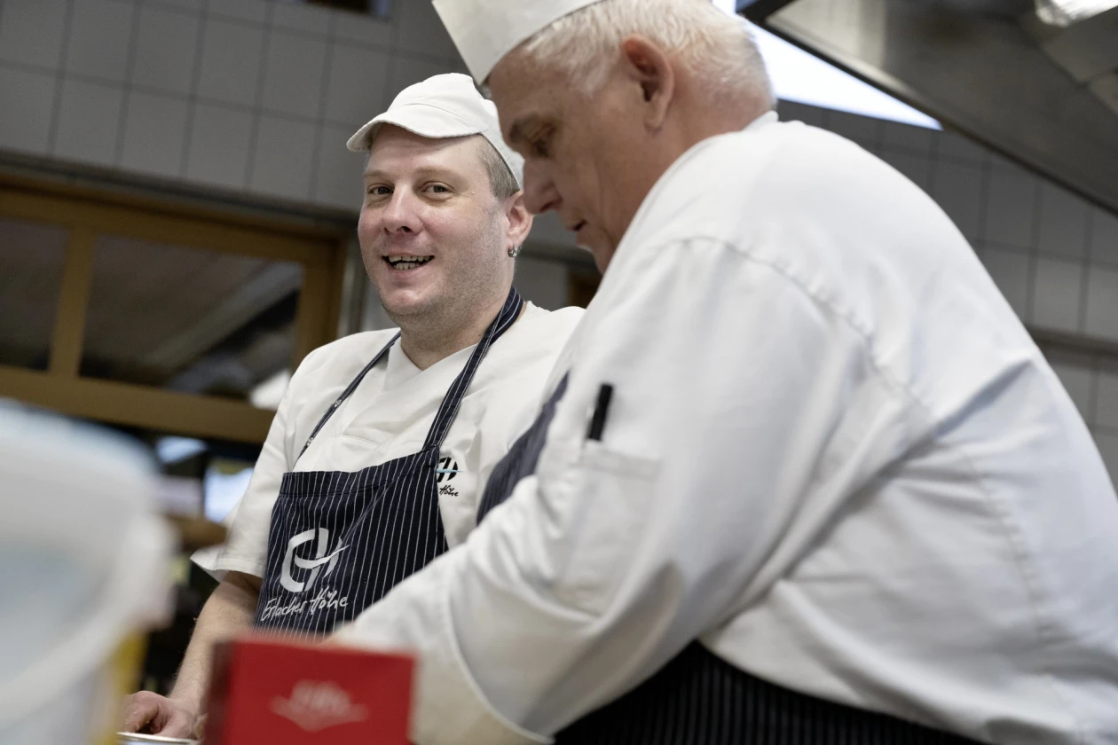 Zwei M&auml;nner in wei&szlig;er Berufskleidung stehen l&auml;chelnd in einer K&uuml;che. Beide tragen gestreifte Sch&uuml;rzen, einer eine wei&szlig;e M&uuml;tze der andere eine Kochhaube