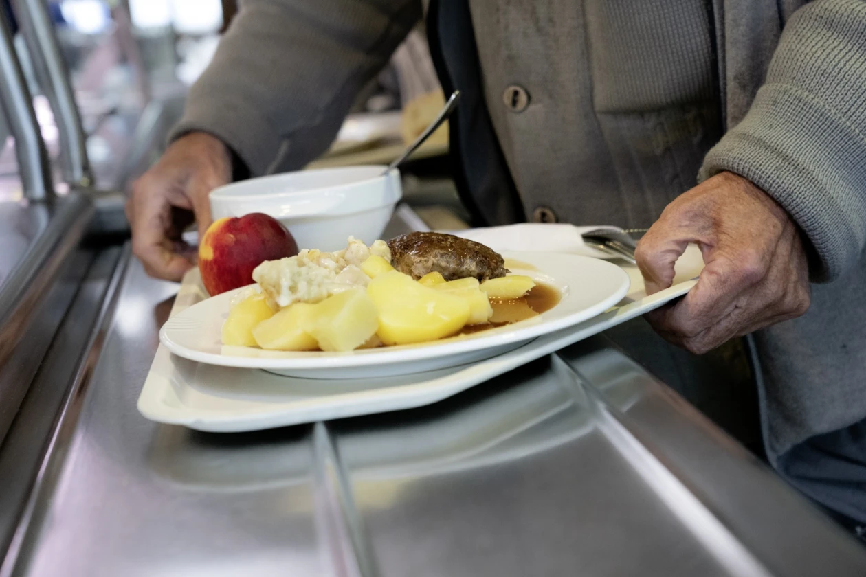 Ein Mann h&auml;lt in einer Kantine ein Tablett mit seiner Mahlzeit in der Hand. Auf dem Teller liegen Salzkartoffeln , ein Hackfleischk&uuml;chle, etwas Gem&uuml;se und daneben ein roter Apfel.