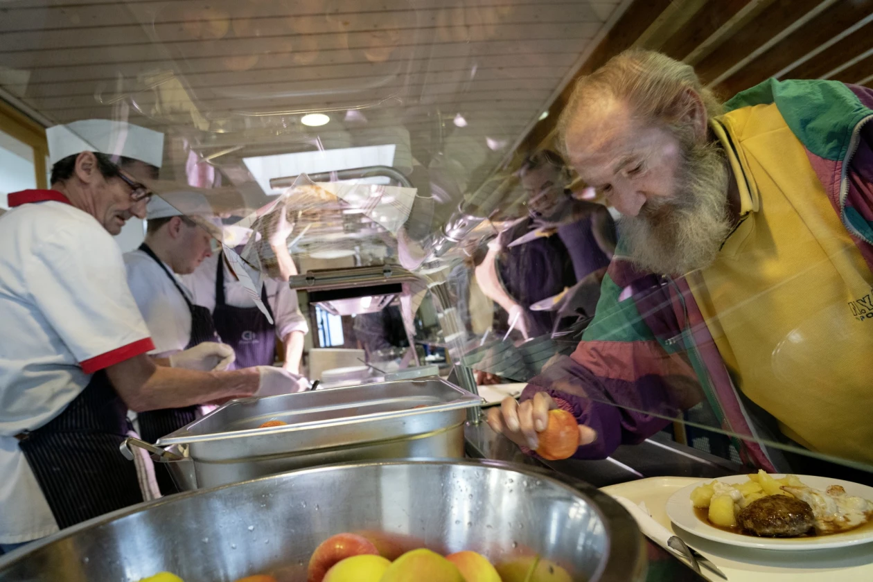 Drei  K&ouml;che in wei&szlig;er Kleidung mit schwarzen Sch&uuml;rzen stehen an der Ausgabetheke einer K&uuml;che. Ein &auml;lterer Mann mit Bart, gelbem Pullover und bunter Jacke hat sein Tablett vor sich und nimmt sich aus einer Edelstahlsch&uuml;ssel einen Apfel.