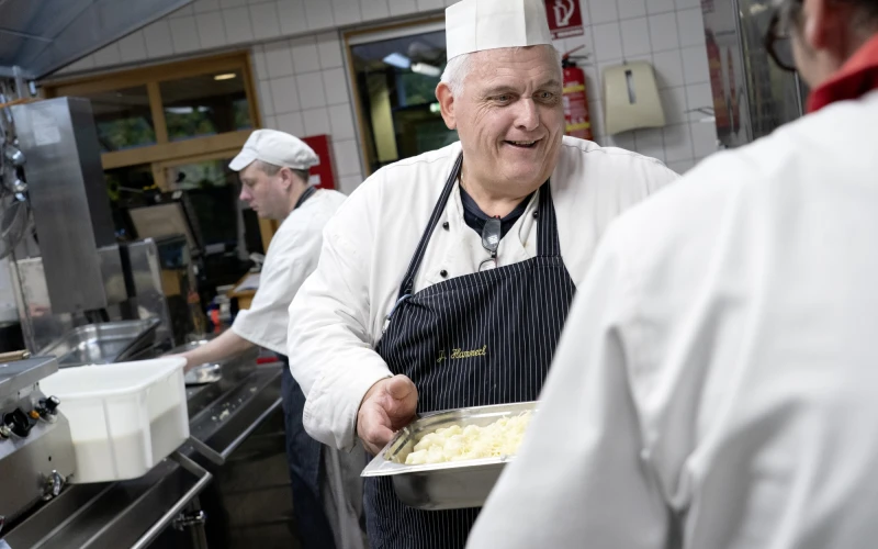 &Auml;lterer Koch in wei&szlig;er Kochjacke und gestreifter Sch&uuml;rze in einer K&uuml;che. L&auml;chelnd h&auml;lt er eine gro&szlig;e Auflaufform. Im Hintergrund sind weitere K&ouml;che bei der Arbeit zu sehen.