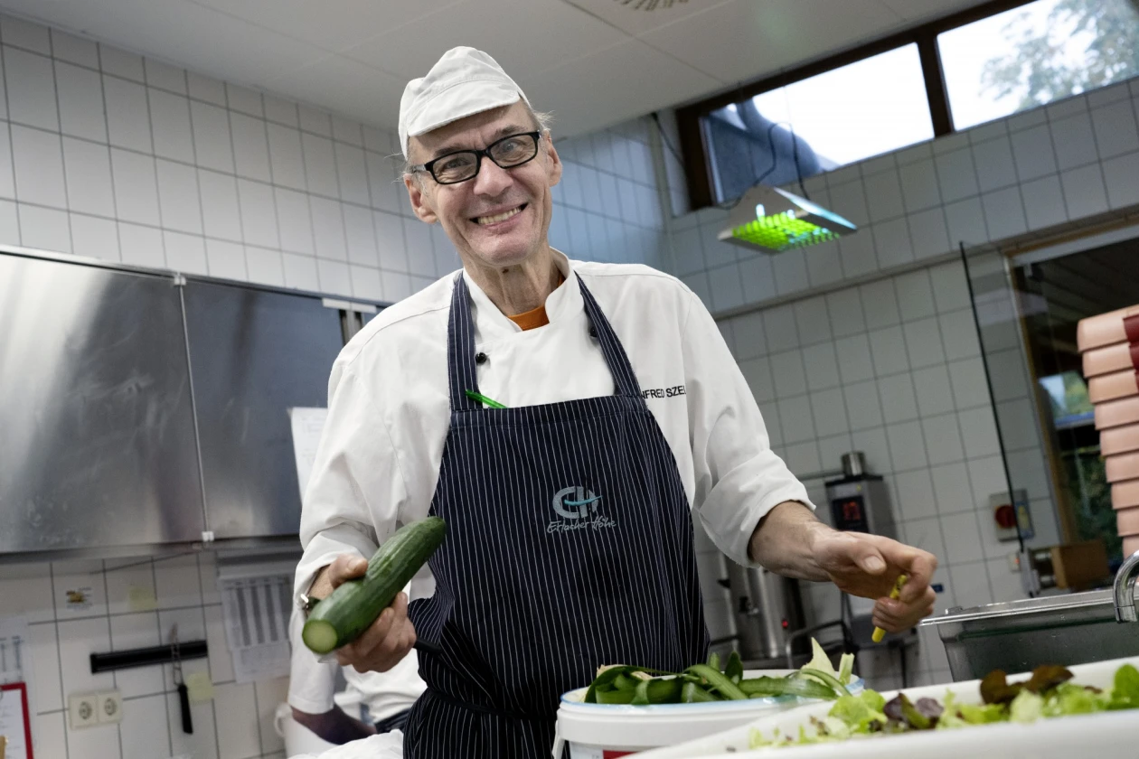 Ein Mann in wei&szlig;er Kochkleidung mit gestreifter Sch&uuml;rze steht l&auml;chelnd in einer K&uuml;che und h&auml;lt eine Gurke in der rechten Hand. Auf dem Tisch vor ihm stehen zwei Beh&auml;lter, einer davon mit gr&uuml;nem Salat.