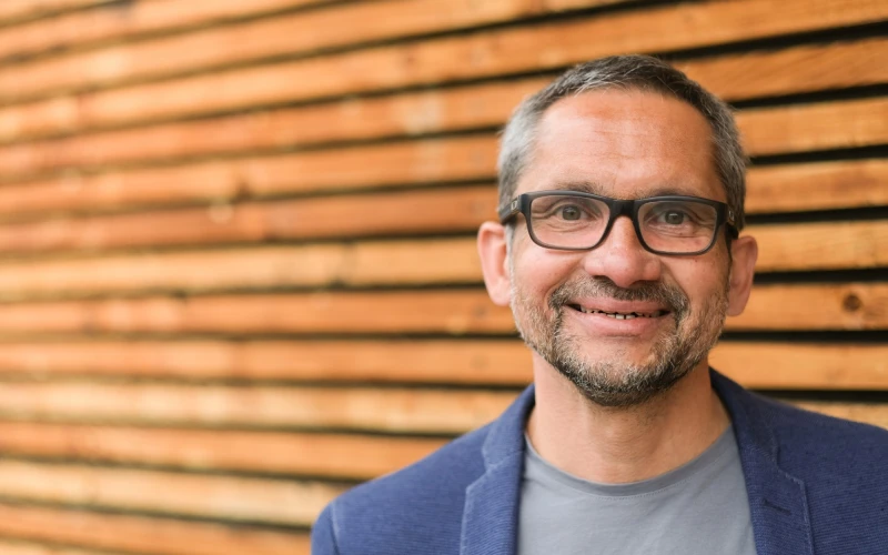 Ein Mann mit Brille und Bart l&auml;chelt freundlich in die Kamera. Er tr&auml;gt ein blaues Sakko und ein graues T-Shirt und steht vor einer Holzwand mit horizontalen Lamellen.