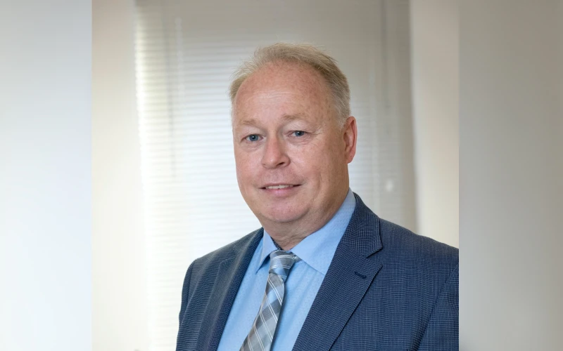 Ein Mann mit dunkelblauem Anzug und gestreifter Krawatte, steht l&auml;chelnd vor einem hellen Hintergrund. Er steht neben einem Computertisch.