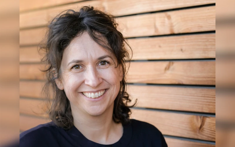 L&auml;chelnde Frau mit schulterlangen, lockigen Haaren vor einer Holzwand. Tr&auml;gt ein dunkles Oberteil.