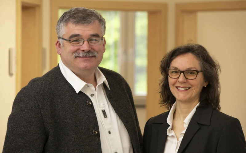 L&auml;chelnder Mann mit Brille, wei&szlig;em Hemd und grauem Sakko neben einer ebenfalls l&auml;chelnden Frau mit Brille, wei&szlig;er Bluse und schwarzem Blazer, die gemeinsam in einem hellen Raum mit Holzt&uuml;ren und Fenster im Hintergrund stehen.