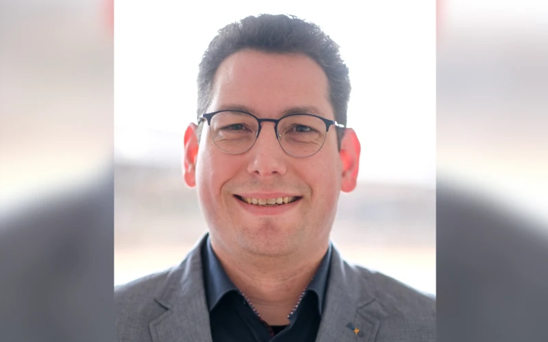 Freundlich l&auml;chelnder Mann mit Brille, grauem Sakko und schwarzem Hemd vor einem unscharfen Hintergrund.