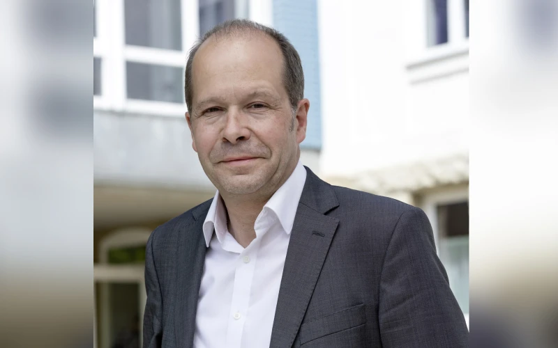 Ein Mann in grauem Sakko und wei&szlig;em, steht vor einem Geb&auml;ude mit Fenstern und gelbem Blumendekor im Hintergrund.