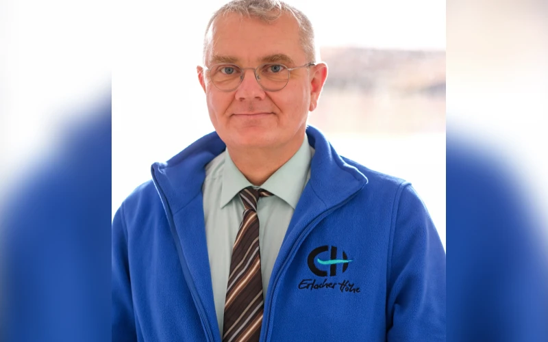 Ein Mann mit Brille tr&auml;gt eine blaue Jacke mit dem EH-Logo und ein mintgr&uuml;nes Hemd mit gestreifter Krawatte. Er l&auml;chelt freundlich in die Kamera und steht vor einem unscharfen Hintergrund.