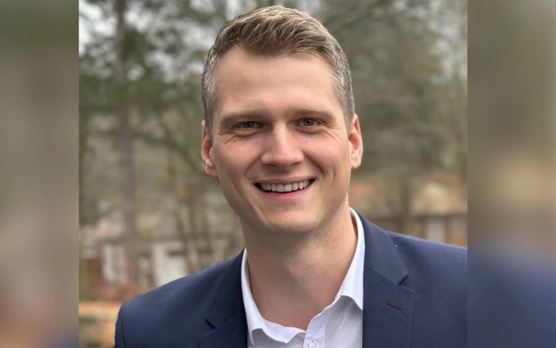 L&auml;chelnder Mann in blauem Sakko und wei&szlig;em Hemd vor unscharfem Hintergrund mit B&auml;umen. Er blickt direkt in die Kamera.