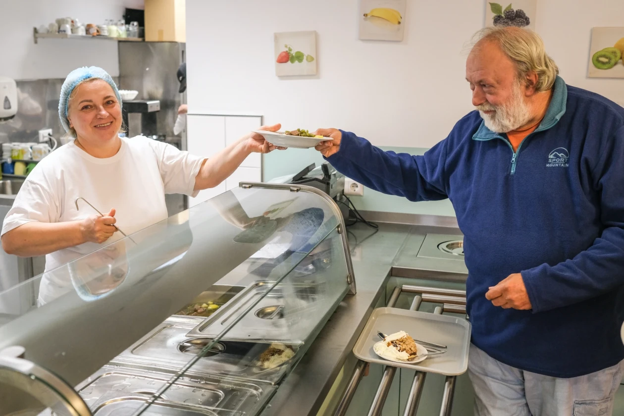 Eine Frau an einer Essenstheke &uuml;bergibt einen Teller mit Essen an einen &auml;lteren Herrn mit Bart und grauen Haaren. In der Hand h&auml;lt sie eine Sch&ouml;pfkelle.