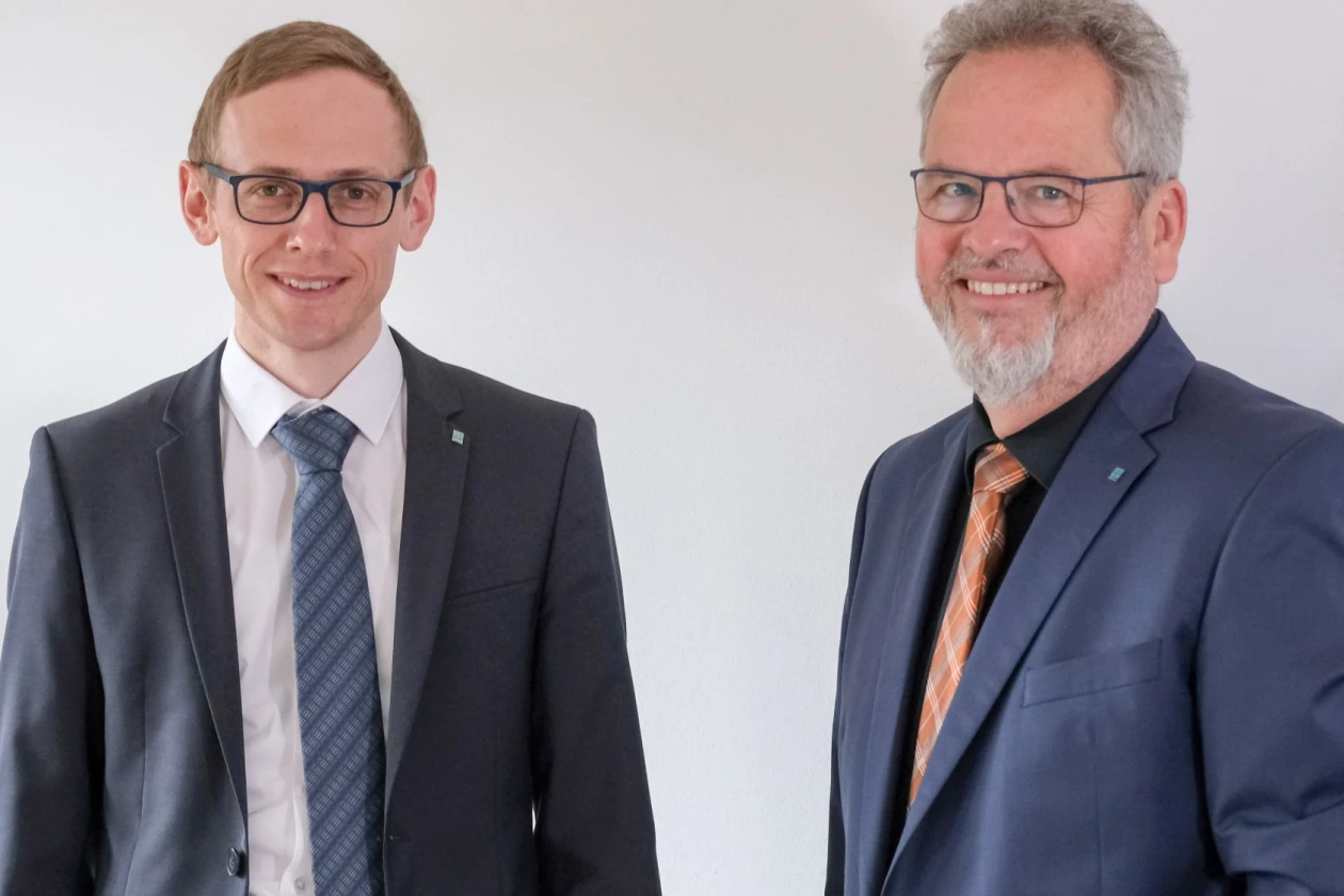 Zwei M&auml;nner in dunklen Anz&uuml;gen, Krawatten und Brillen, stehen vor einem hellen Hintergrund.