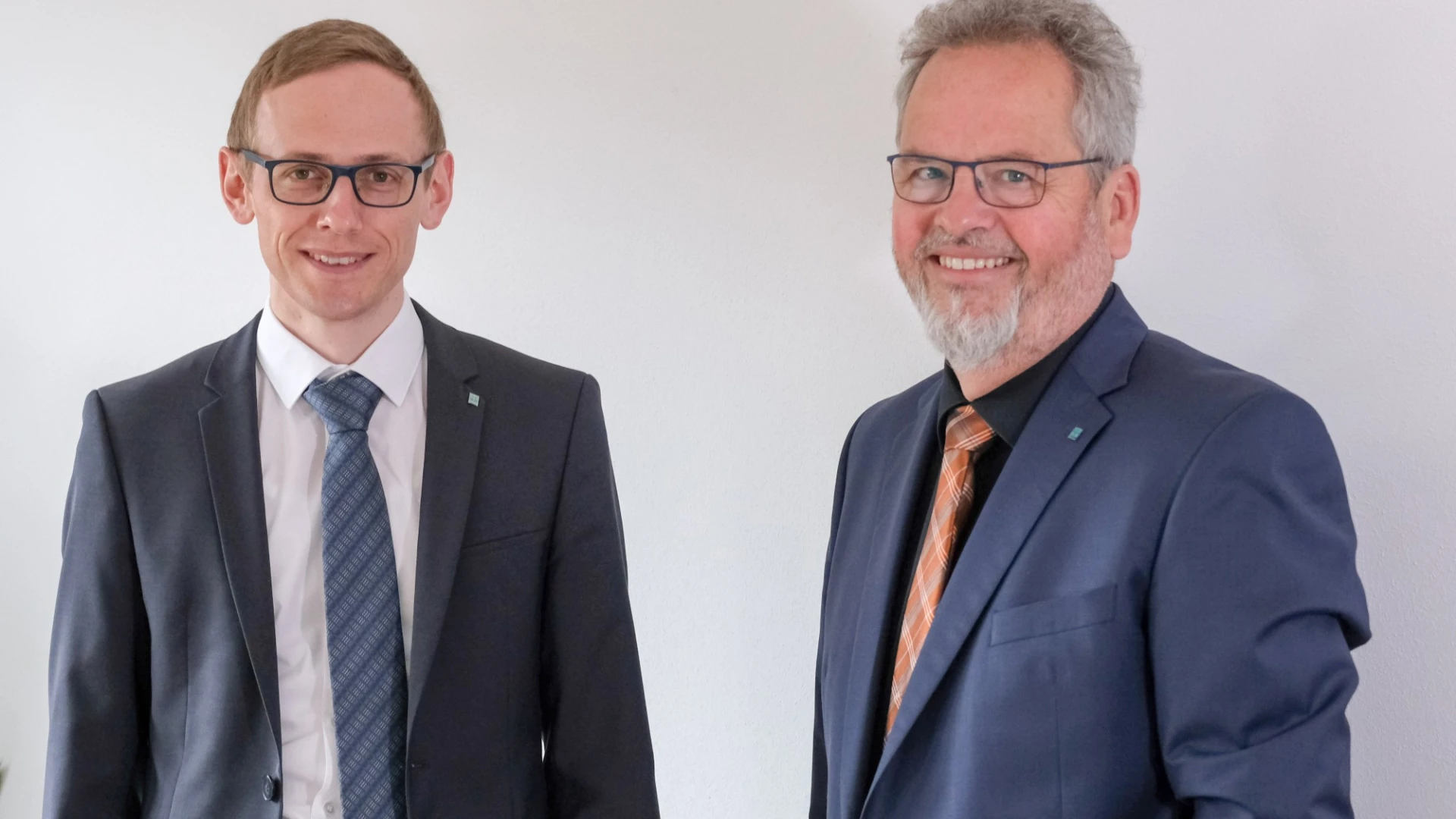 Zwei M&auml;nner in dunklen Anz&uuml;gen, Krawatten und Brillen, stehen vor einem hellen Hintergrund.