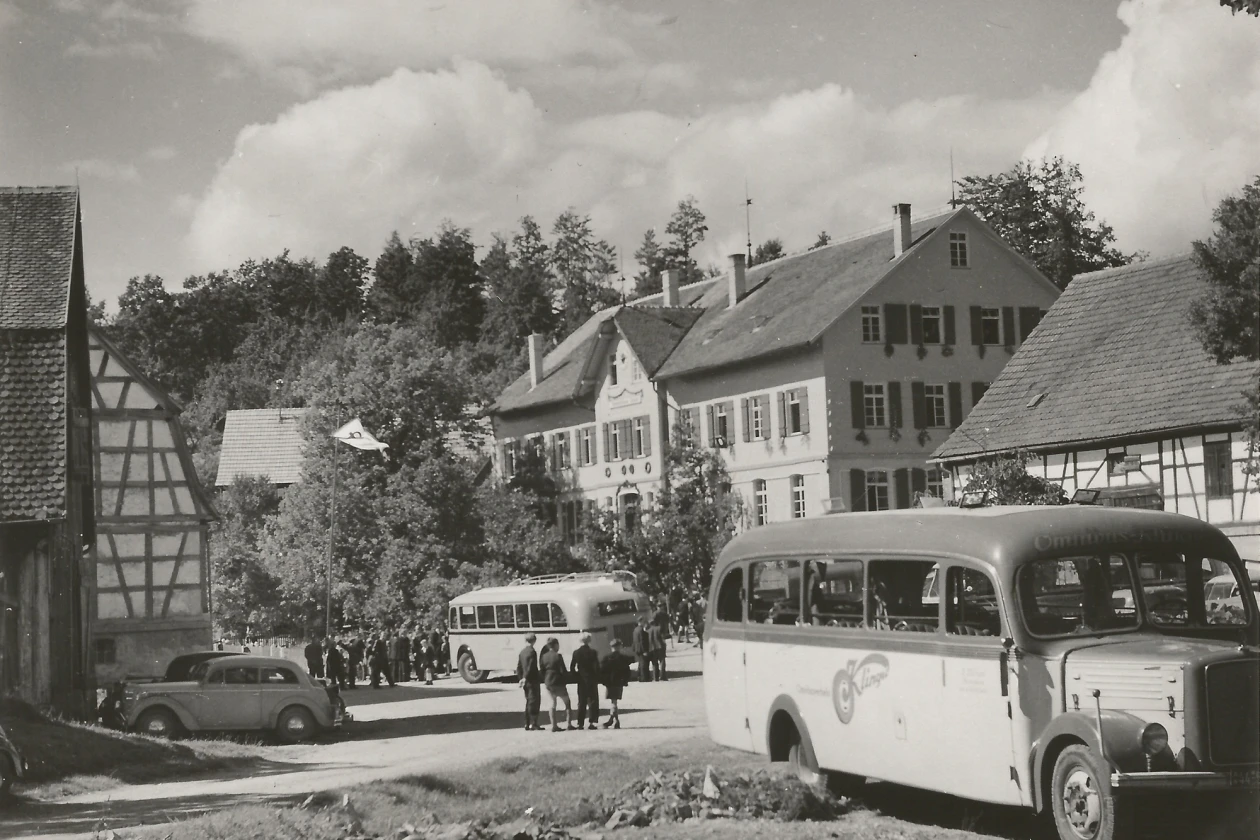 Historische Schwarzwei&szlig;aufnahme eines l&auml;ndlichen Dorfplatzes mit Fachwerkh&auml;usern, B&auml;umen und zwei alten Omnibussen, sowie einem Personenkraftwagen. Im Hintergrund B&auml;ume und bew&ouml;lkter Himmel.