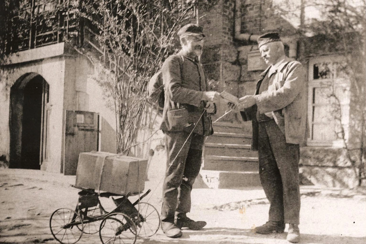 Schwarzwei&szlig;foto von zwei M&auml;nnern, die sich auf einem Gehweg unterhalten. Ein zum Transport umgebauter Kinderwagen ist im Vordergrund. Beide tragen zeitgen&ouml;ssische Kleidung aus der ersten H&auml;lfte des 20. Jahrhunderts und stehen vor einem Backsteingeb&auml;ude mit Treppe.