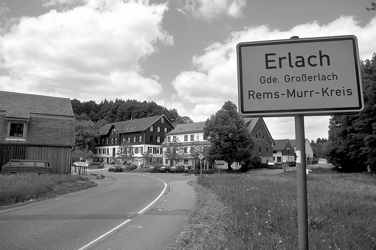 Schwarzwei&szlig;foto vom Ortsschild von Erlach im Rems-Murr-Kreis. Im Hintergrund eine kurvige Landstra&szlig;e und traditionelle Geb&auml;ude im Hintergrund, umgeben von gr&uuml;ner Wiese und Waldh&uuml;gel unter einem bew&ouml;lkten Himmel.