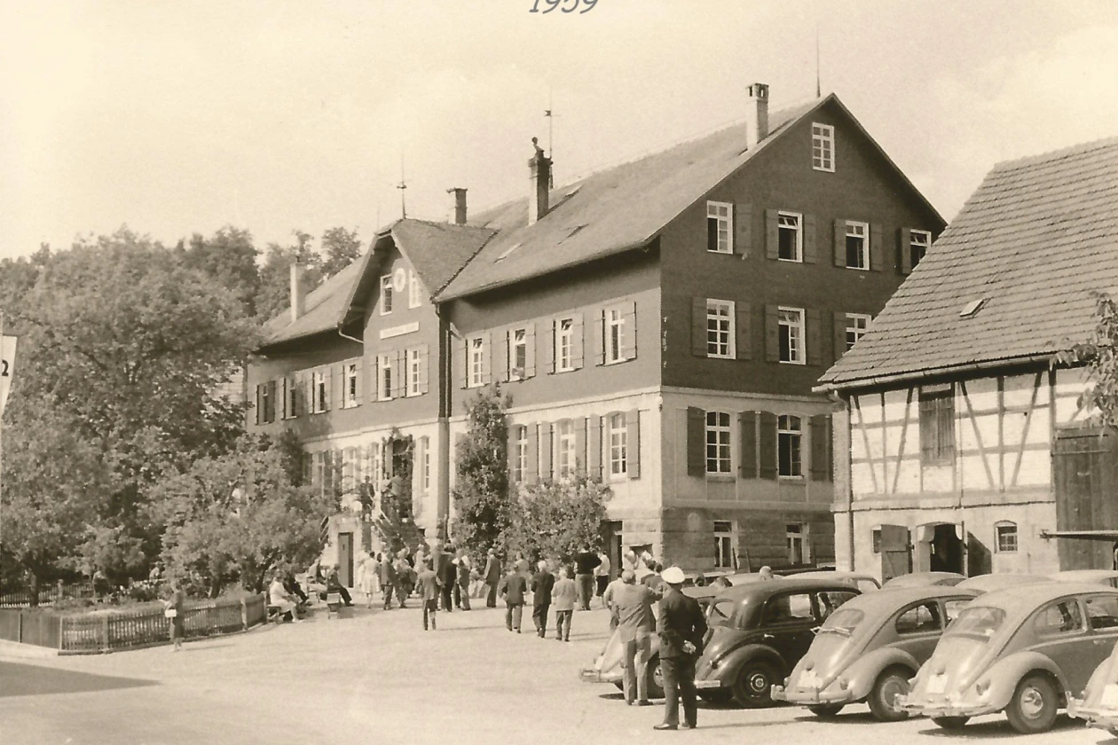 Historische Schwarz-Wei&szlig;-Aufnahme einer Stra&szlig;enszene aus dem Jahr 1959 mit mehreren Geb&auml;uden, parkenden Autos und Personen vor einem mehrst&ouml;ckigen Haus mit Fachwerkelementen und B&auml;umen im Hintergrund.