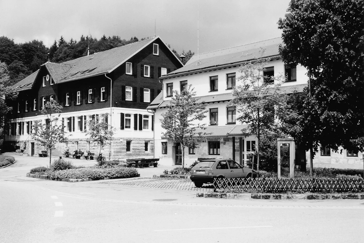 Historisches schwarz-wei&szlig;es Foto eines gro&szlig;en mehrst&ouml;ckigen Geb&auml;udes mit B&auml;umen und einem geparkten Fahrzeug im Vordergrund. Das Geb&auml;ude hat eine dunkle und eine hellere Fassadenseite und liegt in einer l&auml;ndlichen Umgebung mit Waldkulisse im Hintergrund.