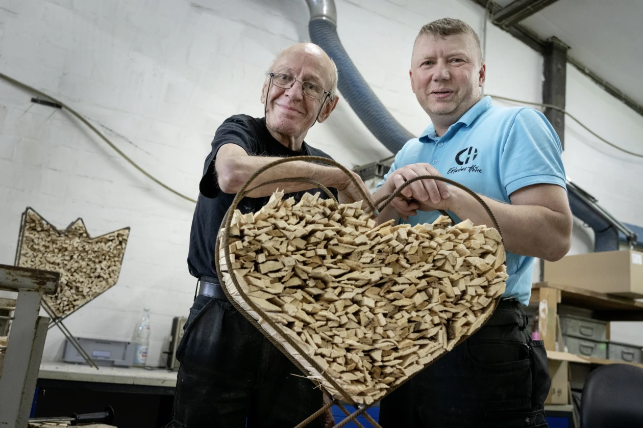 Zwei M&auml;nner halten gemeinsam ein gro&szlig;es, Metallherz mit Anz&uuml;ndholz.