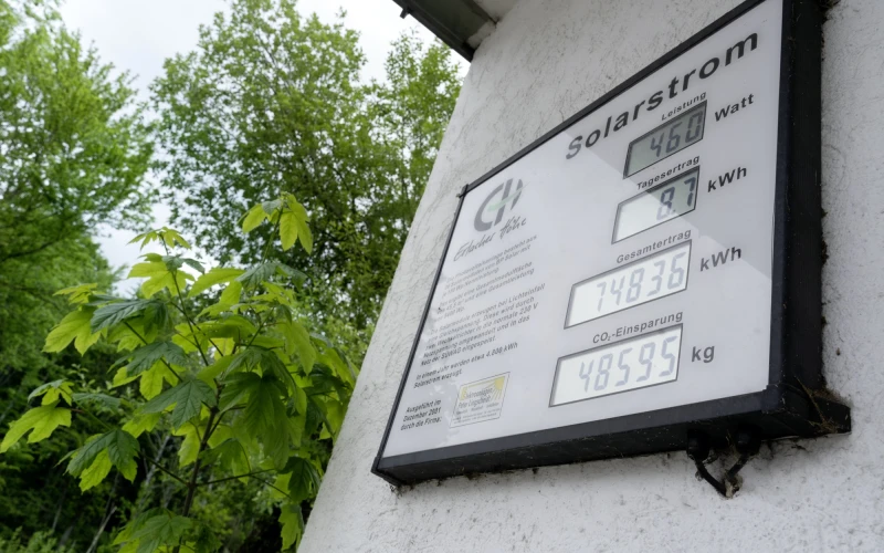 Solarstrom-Messger&auml;t an einer wei&szlig;en Wand, das verschiedene Messwerte wie Watt, kWh und CO2-Einsparung in kg anzeigt, mit gr&uuml;nen Bl&auml;ttern und B&auml;umen im Hintergrund.