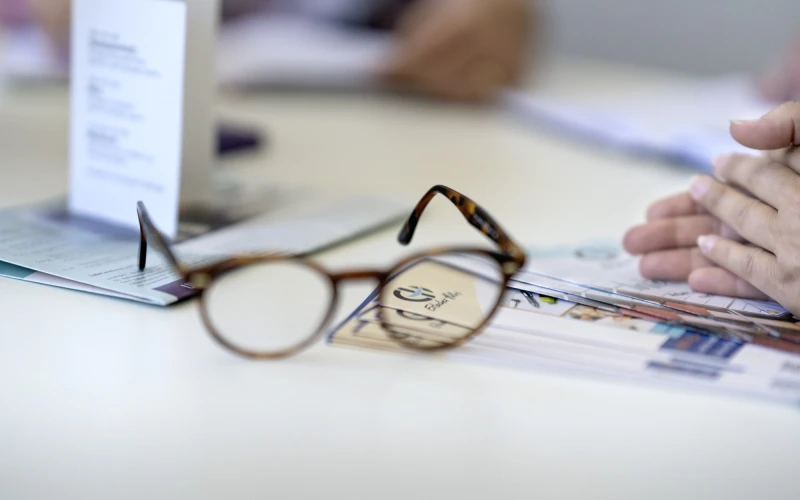 Braune Lesebrille auf einem Papierdokument mit EH Logo und unscharfem Hintergrund, auf dem eine Hand teilweise zu sehen ist. Die Brille liegt leicht schr&auml;g und reflektiert Licht auf einem Arbeitstisch.